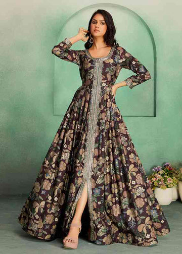 A woman models a long, floor-length printed gown with three-quarter sleeves, a scoop neckline, and a high front slit. The dress features a dark brown base with a multicolor floral and paisley pattern in beige, green, and blue, accented by a silver embroidered border down the front center.