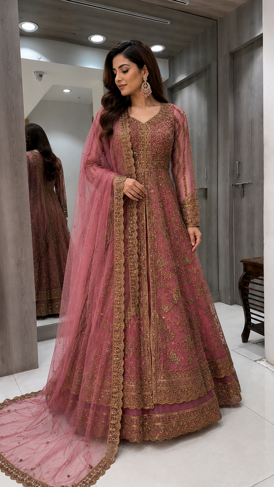 A woman models a traditional Indian anarkali gown in dusty rose pink with intricate gold embroidery, paired with a matching sheer dupatta, in what appears to be a fitting room or boutique.