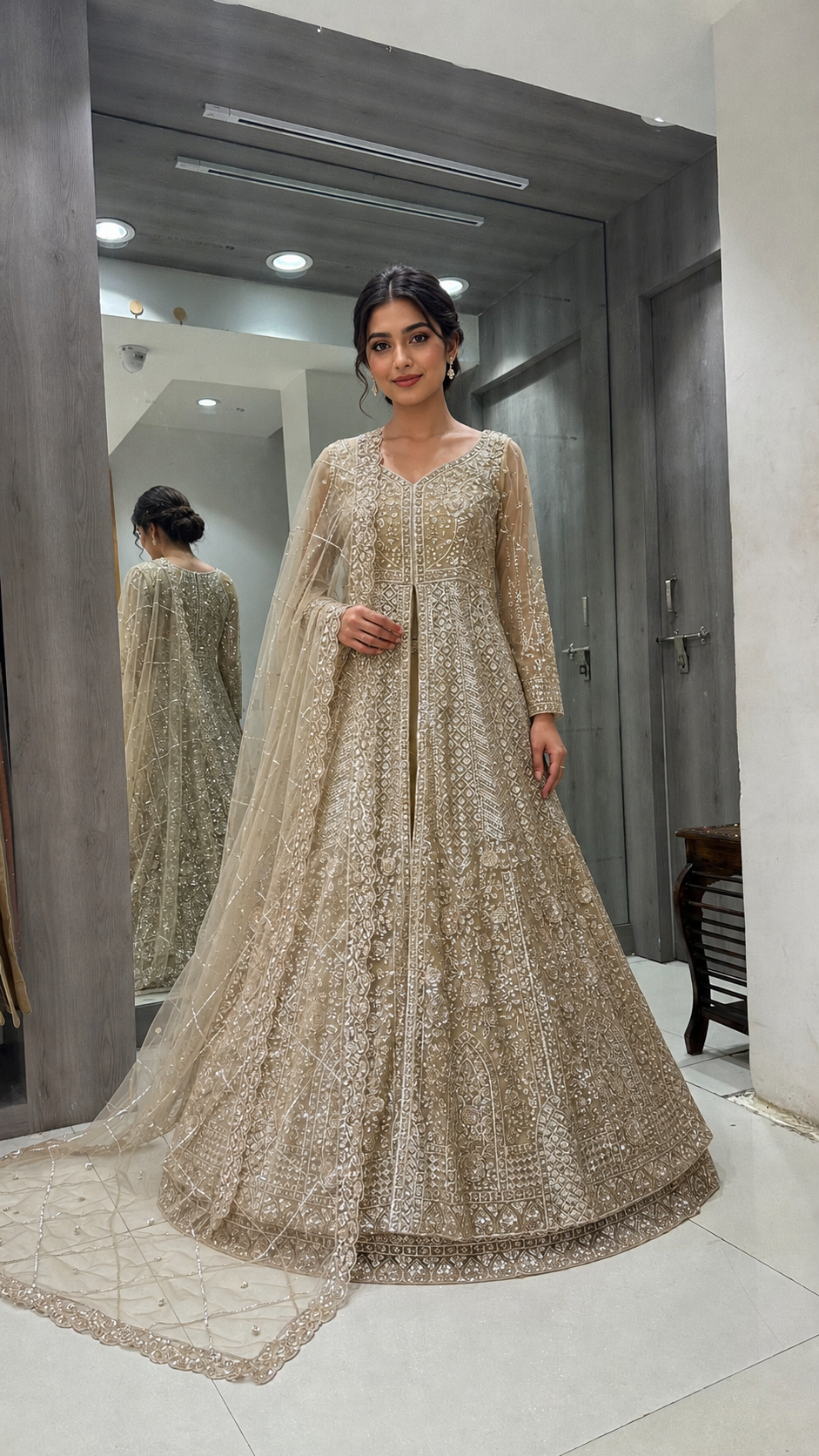 A woman stands indoors wearing a heavily embellished beige/ivory anarkali-style gown with a matching sheer dupatta. The dress features dense gold and silver embroidery and beadwork across the full skirt and sleeves.