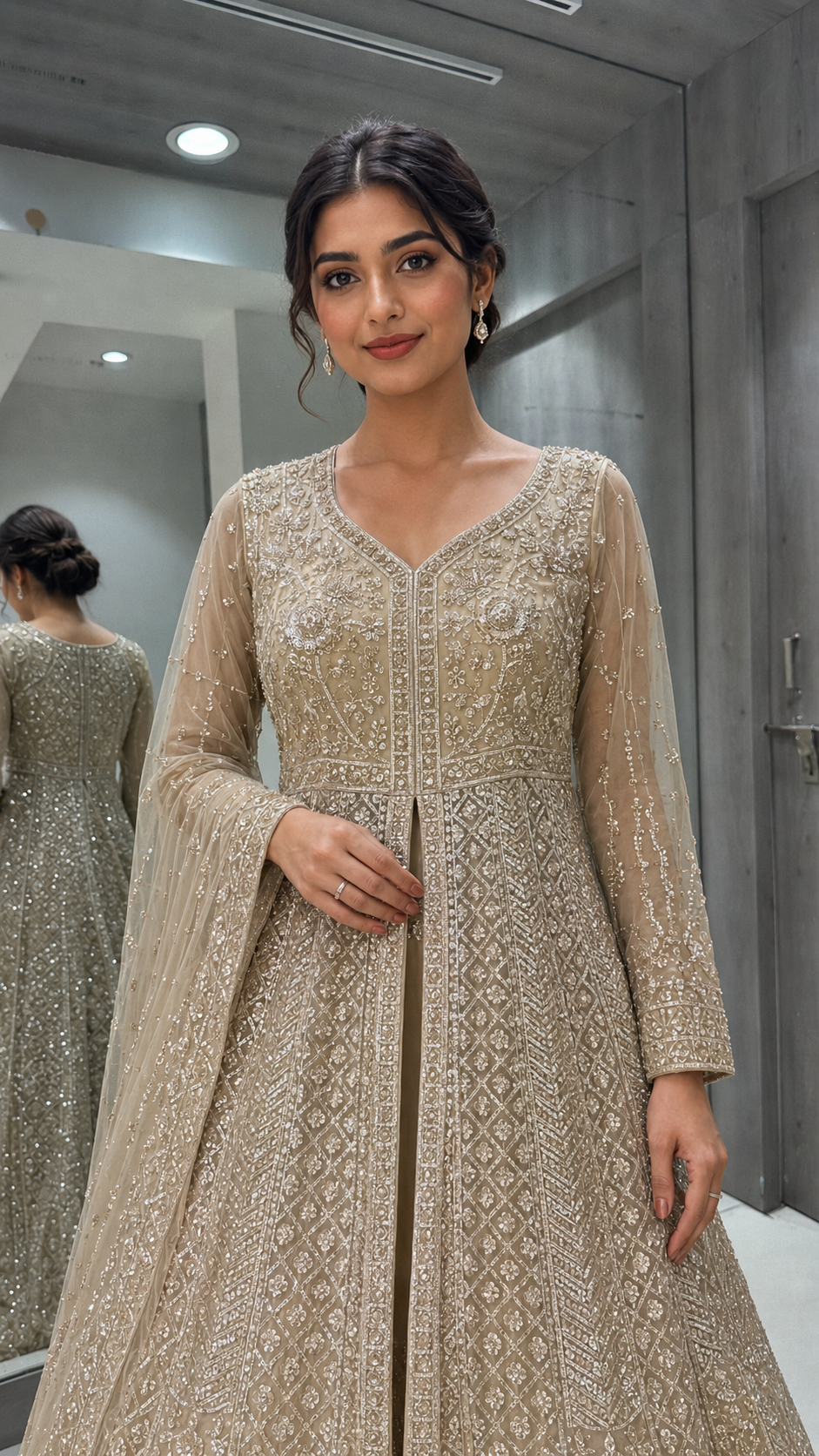 A woman wearing a beige embellished anarkali-style dress with intricate embroidery and a sheer draped sleeve, standing in a fitting room with a mirror behind her showing the back of the dress.