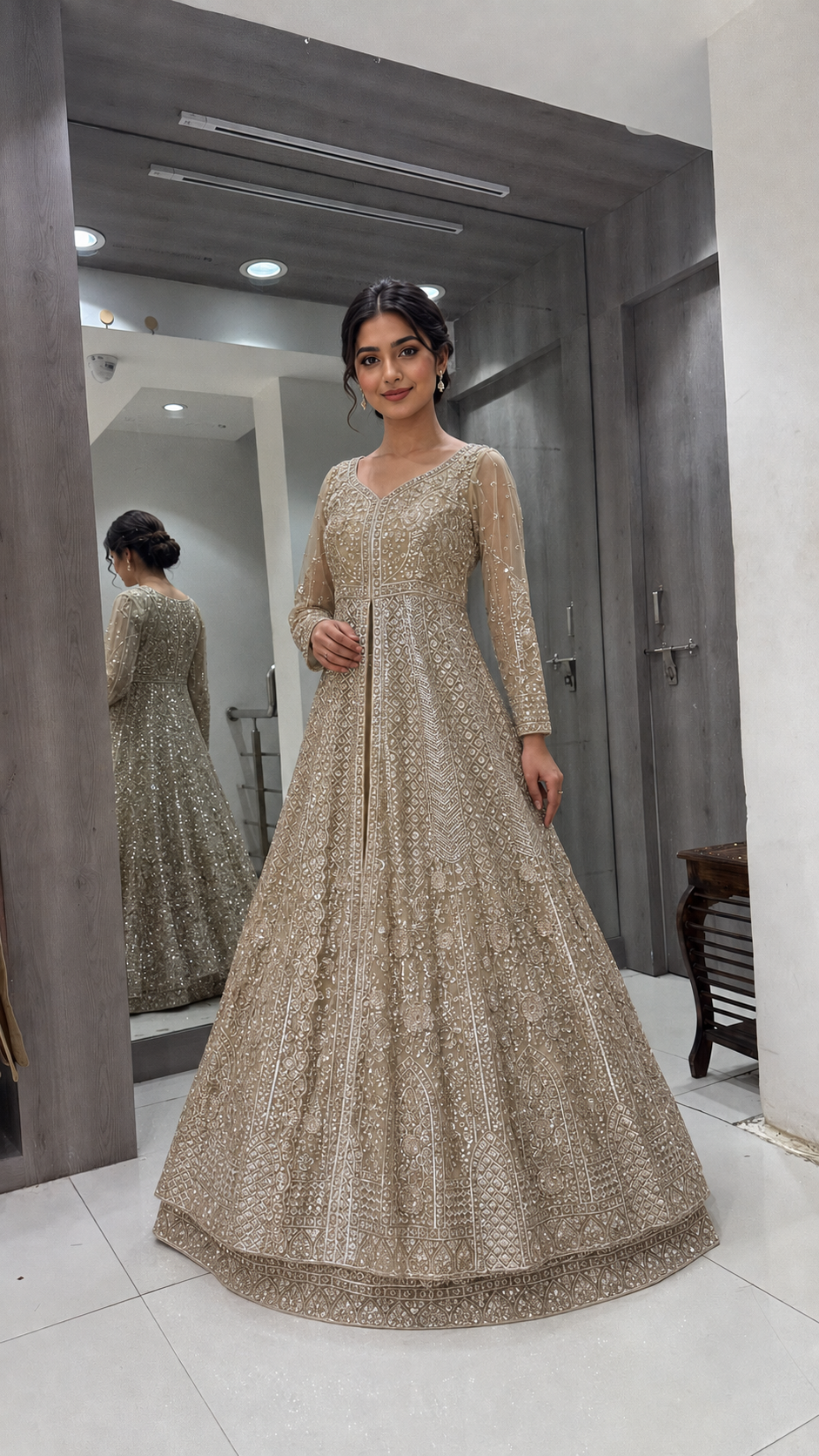 A woman stands in a fitting room wearing a floor-length beige embellished gown with long sleeves and a fitted bodice. A mirror behind her shows the back of the dress, and the room has gray walls and white tiled floor.
