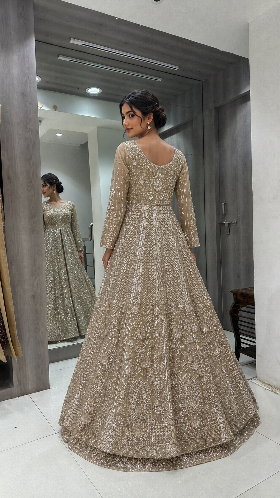 A woman stands indoors wearing a floor-length embellished gown with long sleeves and a fitted bodice, shown from the back. A mirror behind her reflects the front of the dress, and the setting appears to be a boutique or fitting space with gray walls and tiled floor.