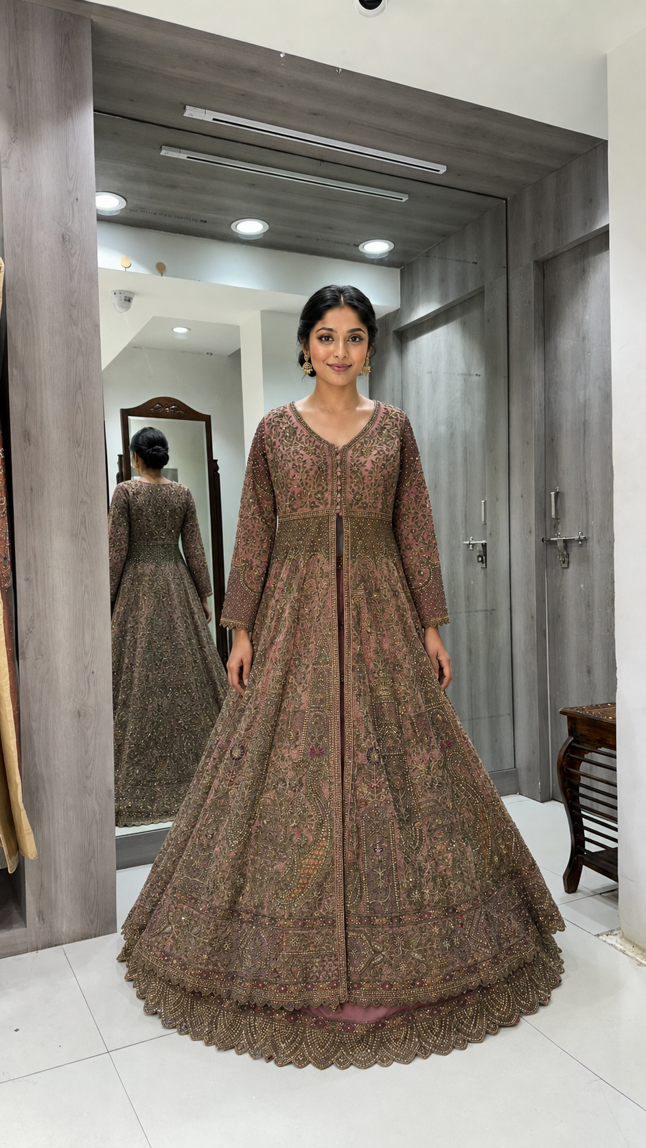 A woman wearing a brown heavily embroidered Anarkali gown stands in a fitting room, with her reflection visible in the mirror behind her.