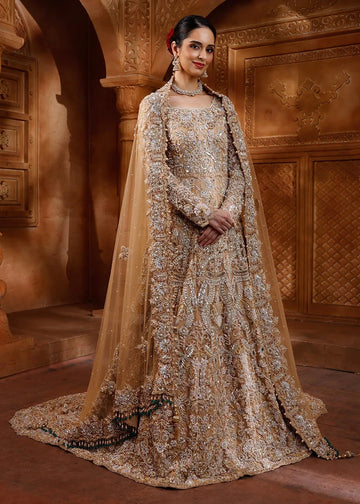 A woman models an ornate beige and gold heavily embroidered traditional gown with a matching sheer dupatta, set against a warm, carved architectural background.